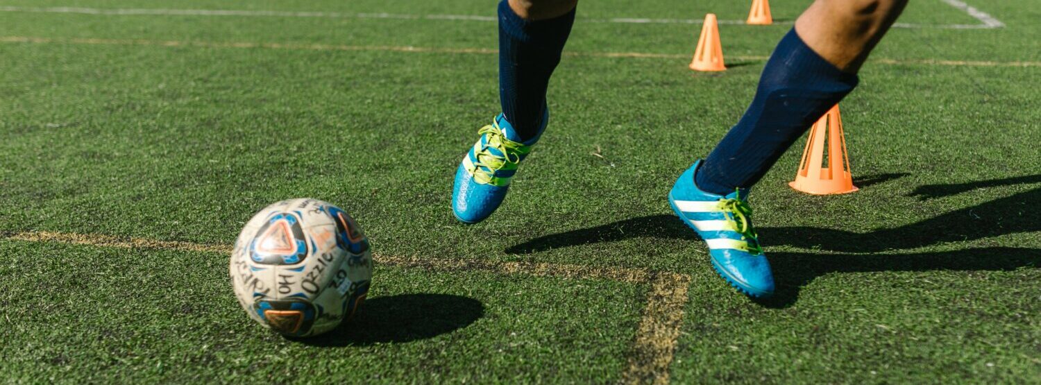 Image of persons legs, wearing football boots on a green pitch. They are dribbling ball around orange cones.