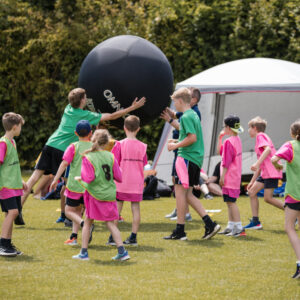 A group of children in pink and green t-shirts. They are playing Kinball. The giant black Kinball is above their heads which some of the children are reaching to touch