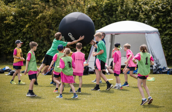 A group of children in pink and green t-shirts. They are playing Kinball. The giant black Kinball is above their heads which some of the children are reaching to touch