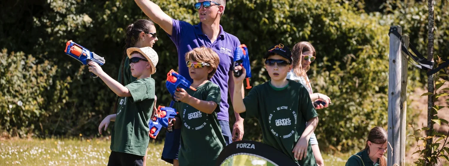 A group of young children engaging in active play. They are using nerf guns on a sunny day on a field.