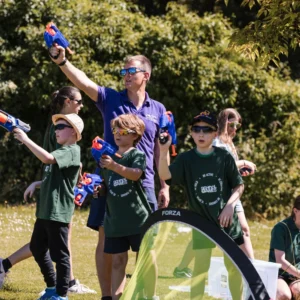 A group of young children engaging in active play. They are using nerf guns on a sunny day on a field.