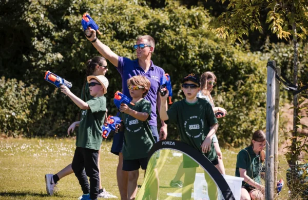 A group of young children engaging in active play. They are using nerf guns on a sunny day on a field.