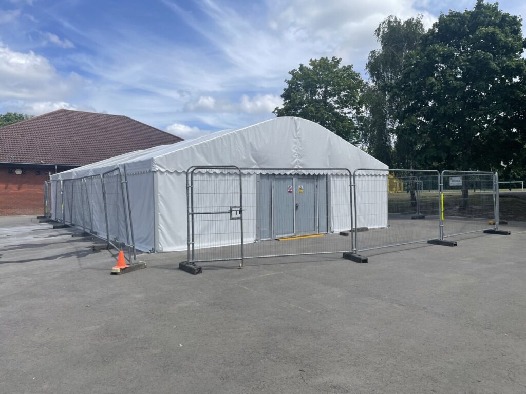 The marquee used to house the pop up pool on a playground. There is security fencing around the marquee