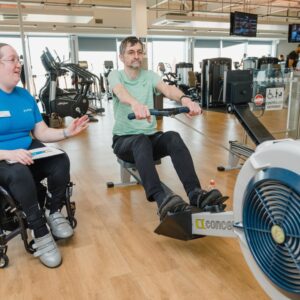 A female gym instructor in a wheelchair overseeing a man on a rowing machine.