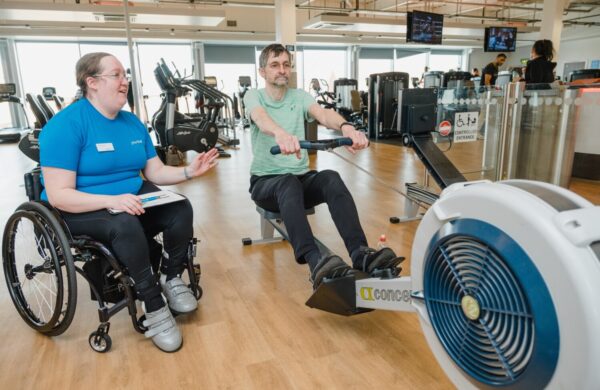 A female gym instructor in a wheelchair overseeing a man on a rowing machine.