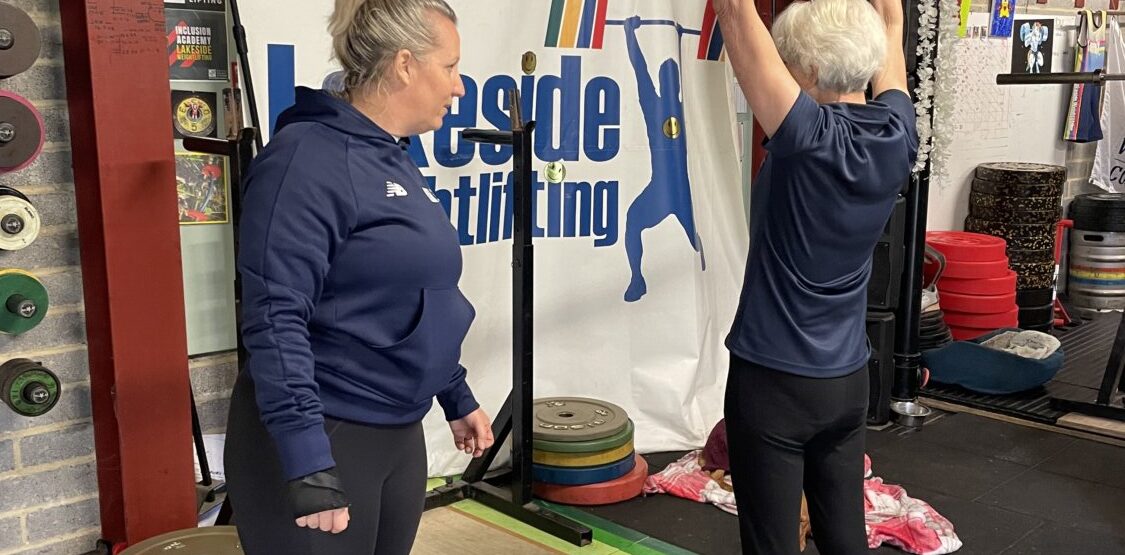 An older lady lifting a bar with weights either end above her head. She is in a gym and being instructed by a coach.