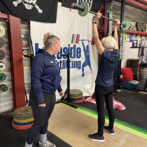 An older lady lifting a bar with weights either end above her head. She is in a gym and being instructed by a coach.