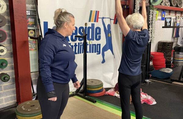 An older lady lifting a bar with weights either end above her head. She is in a gym and being instructed by a coach.